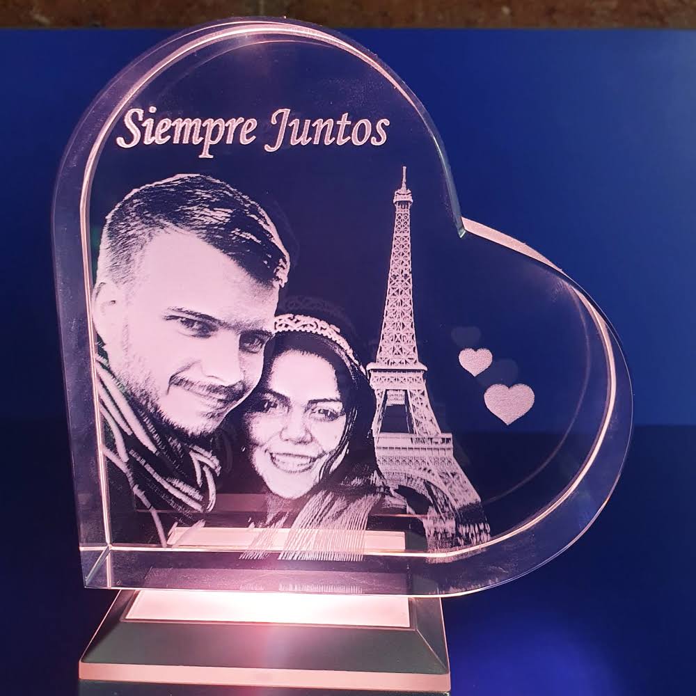 fotocristal-corazon-grabado-siempre-juntos Un bloque de cristal en forma de corazón grabado con láser. En el interior hay un grabado en 3D de una pareja sonriente, la Torre Eiffel y dos corazones flotantes. El texto "Siempre Juntos" está grabado sobre ellos. El cristal está iluminado desde abajo por una base metálica que brilla en color rosa/púrpura, y el fondo es de color azul marino intenso